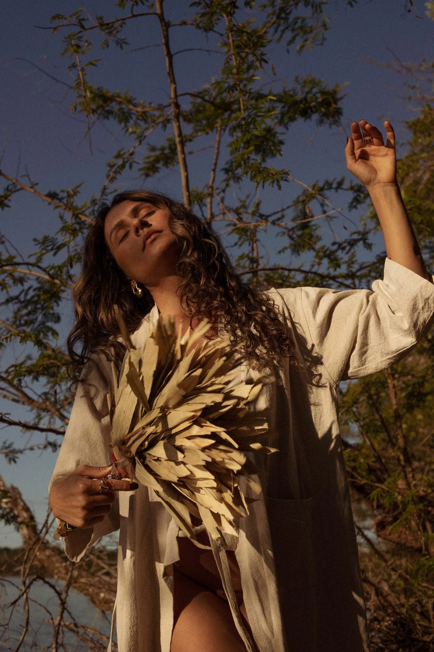 Kalina Cunha na floresta ao entardecer, braços erguidos, banhada por luz dourada